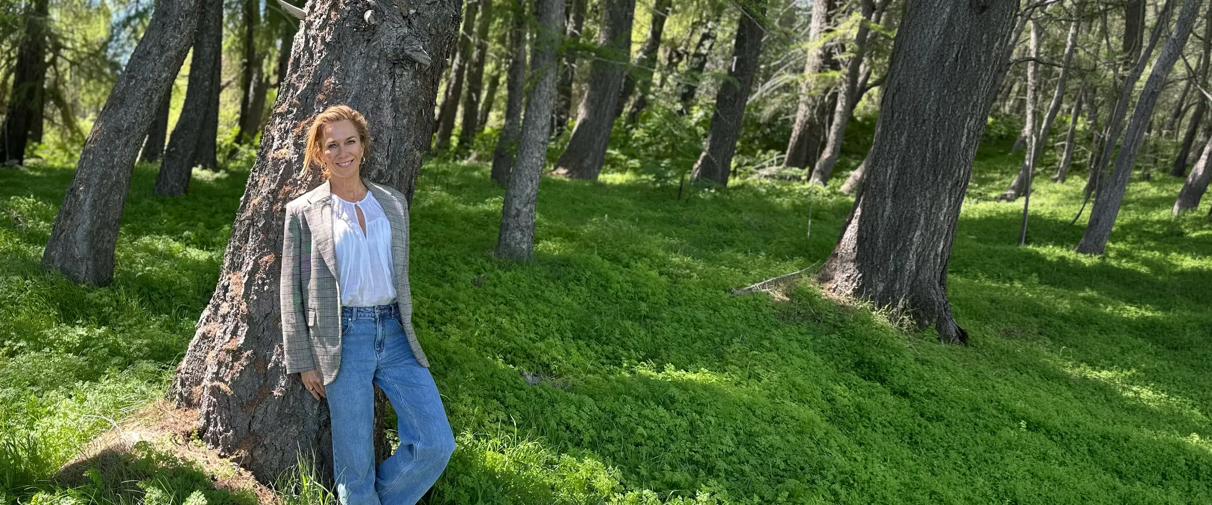 Charlotte, a Hey Human member, standing in a sunlit forest leaning against a tree, smiling in a checked blazer